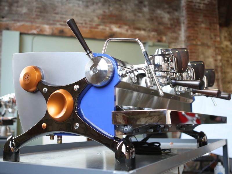 This image is a front-side view of the Slayer Espresso custom machine with custom painted side panels, black nickle plated x-legs, custom painted hubs and accents in Ziricote wood, 3 groups at traditional height with manual dosing controls.